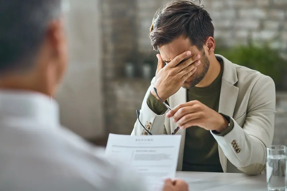 Un hombre vestido con americana se sienta a una mesa, se cubre la cara con una mano y sujeta unas gafas con la otra, parece estresado -posiblemente se enfrenta a un conflicto laboral con su empresa o incluso a un despido improcedente- mientras otra persona revisa un documento en primer plano.