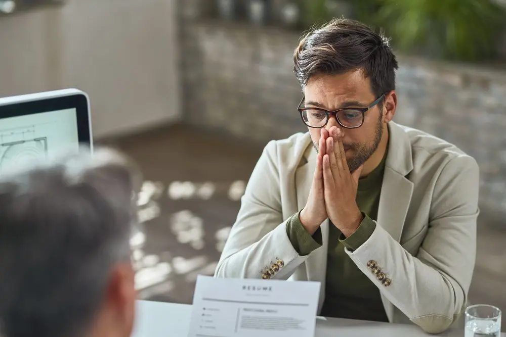Un hombre con gafas y americana beige se sienta a una mesa con las manos juntas cerca de la cara, parece ansioso por algún conflicto laboral con su empresa; en primer plano se ven un documento y otra persona.