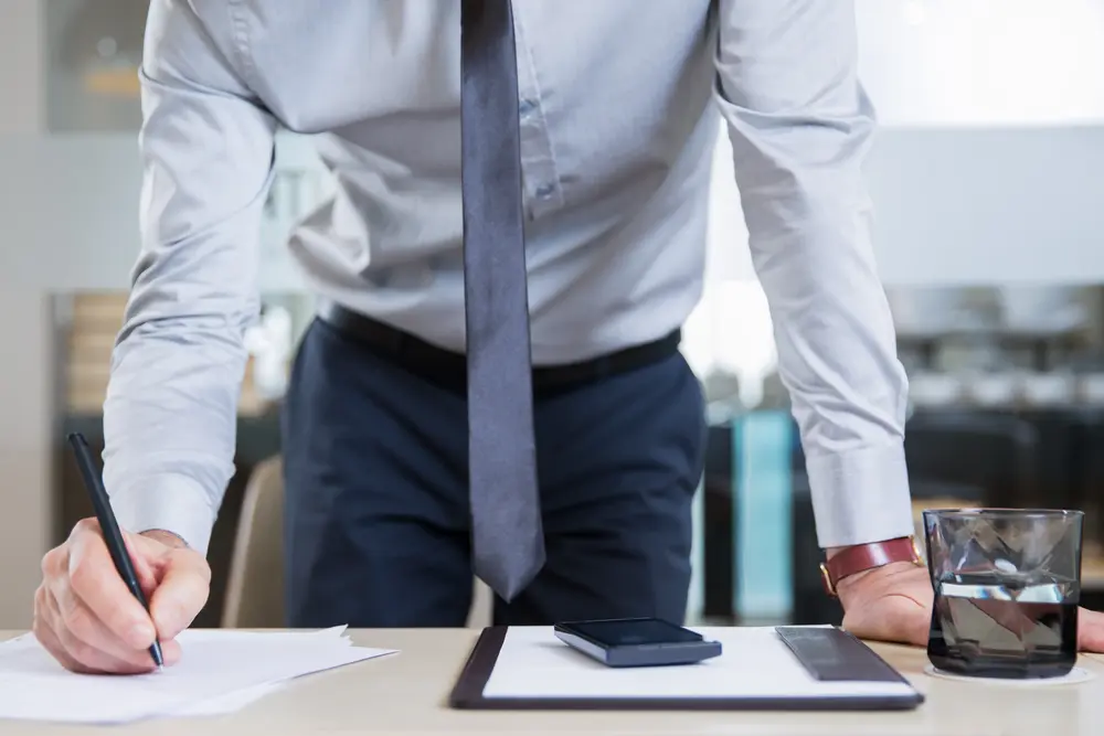 Una persona en traje de negocios se inclina sobre un escritorio, escribiendo en un papel. Sobre el escritorio hay un smartphone, un bloc de notas y un vaso de agua, lo que sugiere un entorno de oficina profesional centrado en la asesoría jurídica.