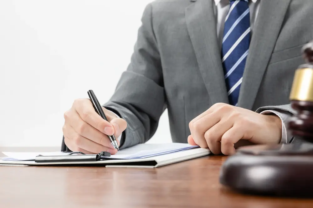 Una persona con traje y corbata a rayas escribe en un cuaderno en un escritorio, con el mazo de un juez visible en primer plano, lo que sugiere que se dedican a la asesoría jurídica.