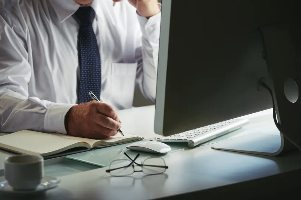 Un hombre con camisa blanca y corbata azul escribe en un cuaderno en un escritorio con un ordenador, un ratón, gafas y una taza, lo que sugiere una oficina o un entorno de trabajo centrado en la asesoría jurídica.