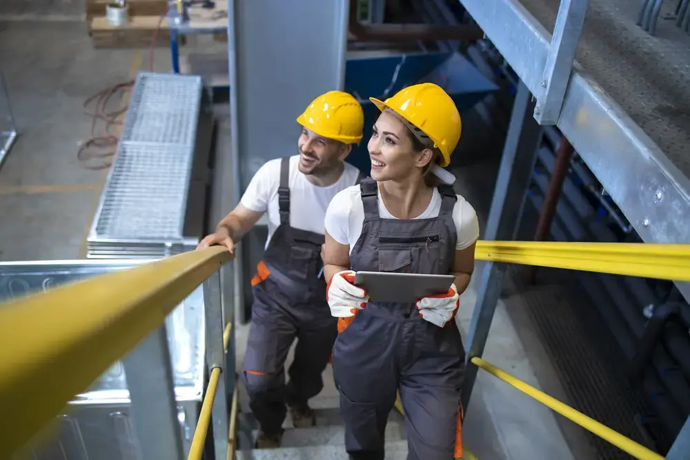 Dos trabajadores de la construcción con cascos amarillos y guantes de seguridad suben las escaleras metálicas de una nave industrial; uno de ellos sostiene una tableta, posiblemente revisando las directrices de asesoría laboral, y ambos sonríen y miran hacia delante.