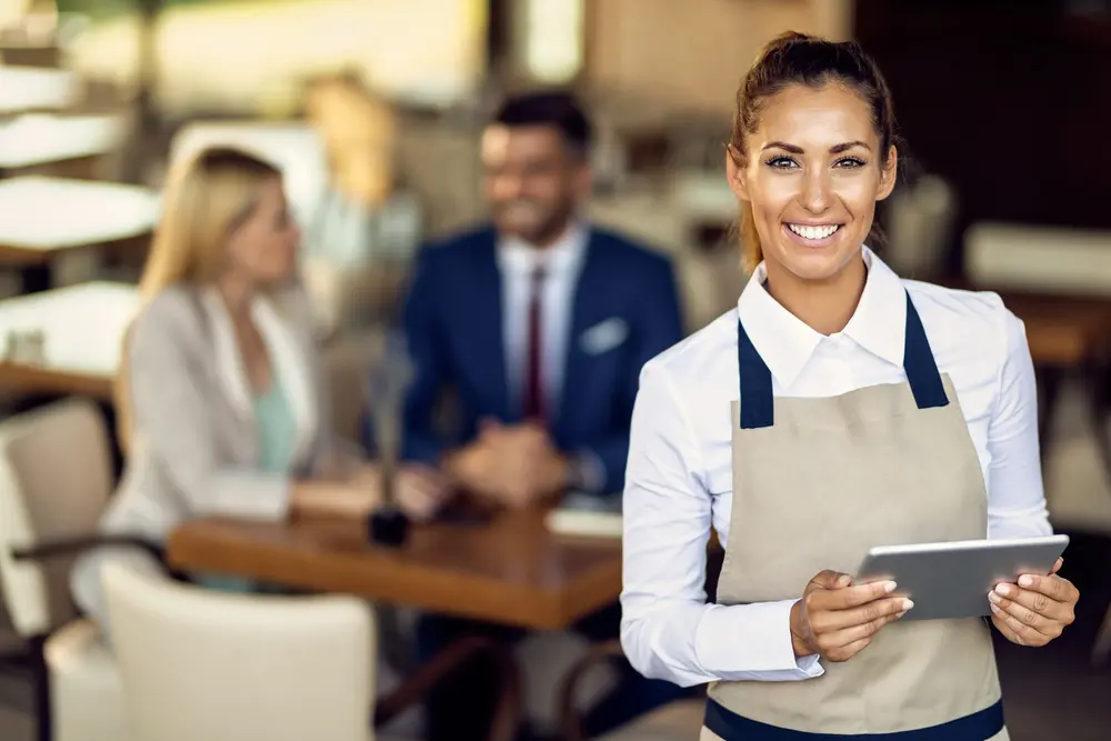 Una sonriente camarera con delantal sostiene una tableta digital en un restaurante, con dos personas en traje de negocios discutiendo asesoría laboral en una mesa en el fondo borroso.