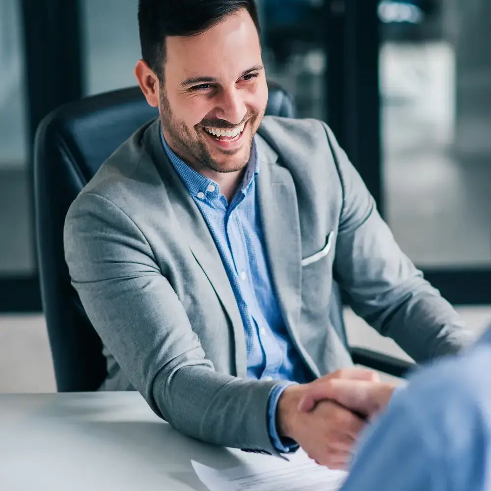 Un hombre sonriente, vestido con americana gris, se sienta ante un escritorio y estrecha la mano de otra persona. Sobre el escritorio se ven papeles y la interacción parece profesional, posiblemente una entrevista de trabajo o una reunión de negocios.