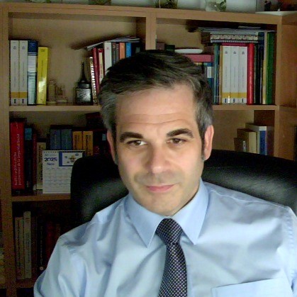 Víctor Pérez González, asesor laboral y Graduado Social Colegiado en Málaga, un hombre de pelo corto y canoso, con camisa azul claro y corbata oscura, se sienta en una silla de oficina frente a una estantería llena de libros y carpetas.